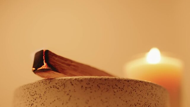 A glowing burning palo santo stick rests on a ceramic holder, with soft candlelight radiating warmth, creating a serene and calming ambiance perfect for relaxation, meditation, or spiritual practices.