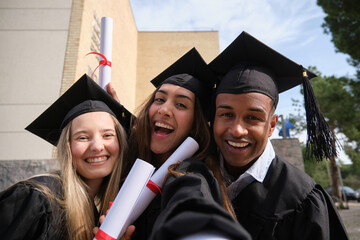 Three cheerful multi ethnic students wearing graduation gowns and caps are taking a selfie holding...