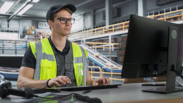 Male Stocking Manager Checking Inventory On Desktop Computer In Warehouse Facility With Automated Conveyor Belt. Logistics Company Employees Carrying Boxes to Package Products And Deliver to Clients.