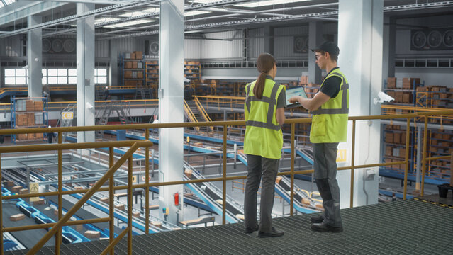 Caucasian Male Logistics Specialist And Female Transportation Expert Talking And Using Laptop In Automated Warehouse Facility With Conveyor Belt. Packaging, Delivering Products to Customers Concept