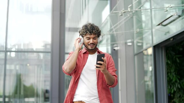 Happy young businessman wearing wireless headphones is enjoying music from his mobile phone while walking on the street near office building. Handsome joyful man is smiling and in a good positive mood - Powered by Adobe