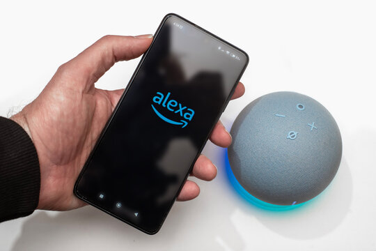 Mano sosteniendo un smartphone con el logo de Alexa junto a un altavoz inteligente Echo Dot sobre un fondo blanco con el anillo de luz azul activado y mostrando la hora