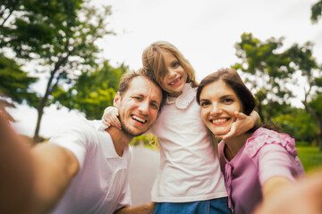 Happy family taking selfie at park. White mother and father taking a photo with daughter with teeth smile.