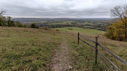 Obraz premium Dirt hiking trail winds down a grassy hillside next to a rustic wooden fence, overlooking a scenic valley under an overcast autumn sky.