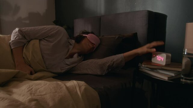 Young woman waking up early in morning, disabling her digital alarm clock and taking off pink sleep mask in cozy bedroom