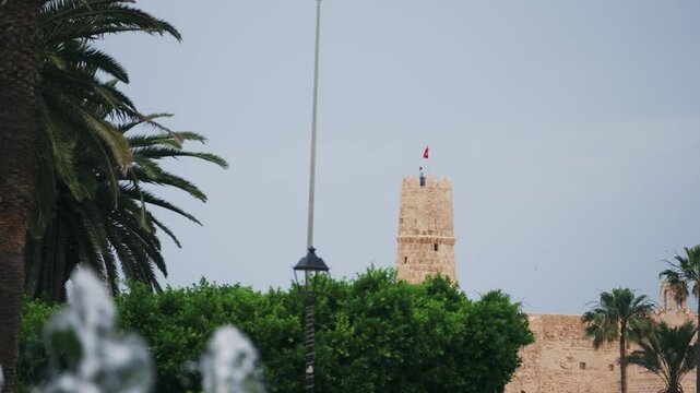 Historic ribat stone tower in Monastir, Tunisia with waving red Tunisian flag surrounded by palm trees and lush greenery