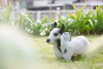 A white cow statue with black horns sits on a lawn with trees and green plants in the background.