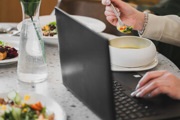 A hand holding a spoon takes soup while typing on a laptop. Plates of food and a glass of water are on the table, showcasing a casual dining and working environment.