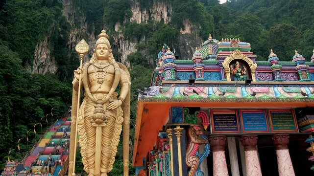 Golden murugan statue greeting visitors near colorful limestone steps leading to sacred hindu temple complex at batu caves, malaysia