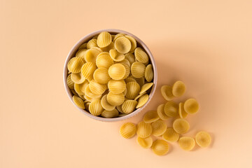 Top view of a porcelain bowl of raw italian durum wheat orecchiette pasta against a light ochre...