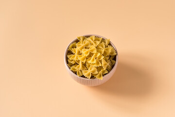 Top view of a bowl of raw italian durum wheat farfalle pasta against a light ochre background .