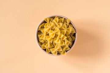 Top view of a bowl of raw italian durum wheat farfalle pasta against a light ochre background .