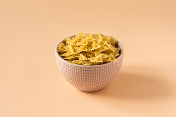 Top view of a bowl of raw italian durum wheat farfalle pasta against a light ochre background .