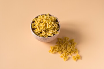 Side view of a porcelain bowl of raw italian durum wheat farfalle pasta against a light ochre...