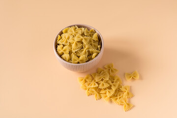 Top view of a porcelain bowl of raw italian durum wheat farfalle pasta against a light ochre...