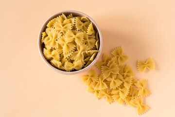 Top view of a porcelain bowl of raw italian durum wheat farfalle pasta against a light ochre...