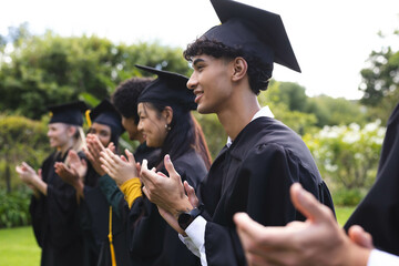 Graduating diverse students in caps and gowns clapping in garden, celebrating achievement