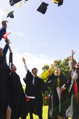 Diverse graduates in caps and gowns joyfully throwing hats in air in garden, copy space