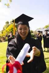 Graduate in cap and gown joyfully reaching for diploma in garden with diverse friends