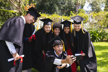 Graduating diverse students celebrating in garden, taking selfie with joyful smiles and diplomas
