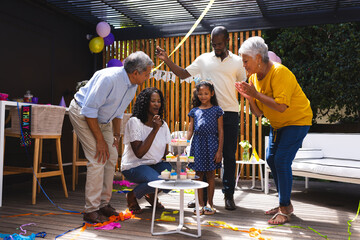 Celebrating birthday in garden, diverse family smiling and clapping around cake and decorations