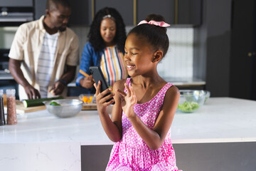In kitchen, girl using smartphone while diverse parents prepare food, at home