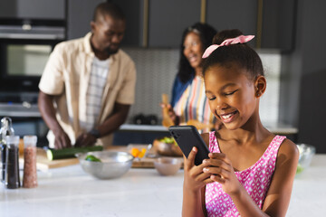 Smiling girl using smartphone in kitchen while diverse family prepares meal together, at home