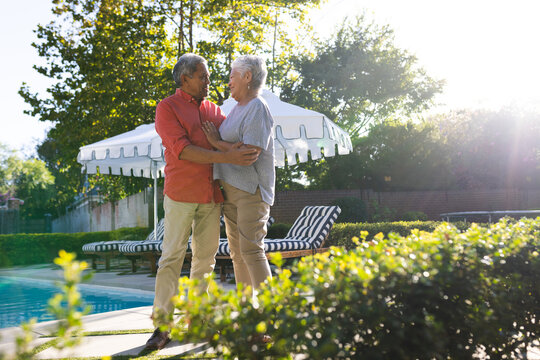 Elderly diverse couple embracing by poolside, enjoying sunny day in garden together, copy space