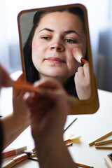 Vertical image of Millennial Caucasian woman doing self make up applying blush on cheeks with make...