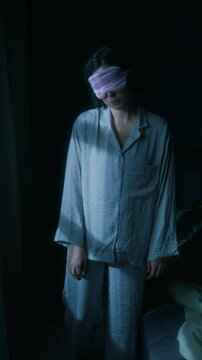 Vertical shot of young woman, wearing pink sleep mask, sleepwalking around bedroom in her pajama at night