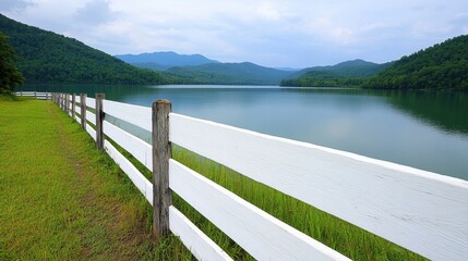 Serene landscape featuring a white wooden fence alongside a calm lake, lush green grass, and a mountain range in the background under a cloudy sky