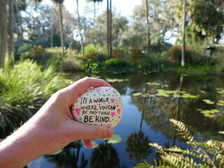 Be kind message painted on kindness rock held in hand in front of tropical water scene
