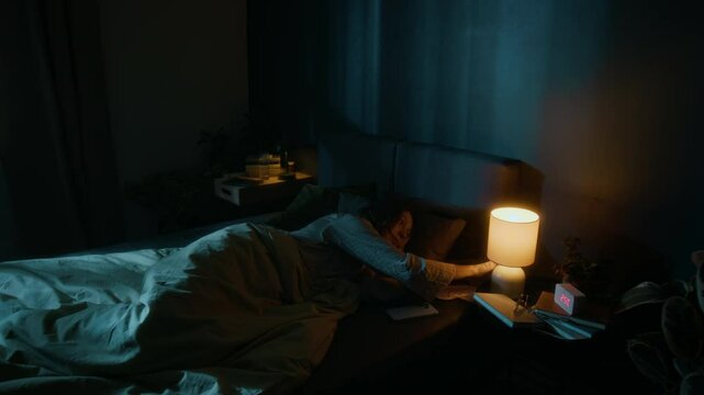 Young woman putting down her smartphone, turning off light on bedside table and falling asleep in bed in modern bedroom