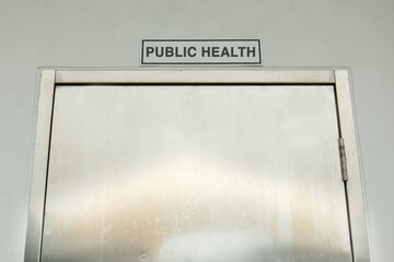 "Public Health" text or word displayed above a steel door on a white wall. A reference to community well-being, healthcare, and disease prevention