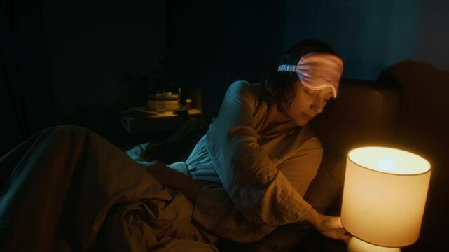 Young woman with dark short hair putting on pink sleep mask and turning off lights at bedside table before sleep at night in modern bedroom