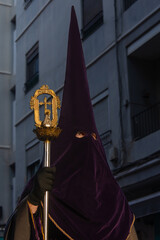 Close-up of the face of a penitent hidden under a high hood, which only reveals his eyes, while he marches in the Holy Week procession and firmly holds a metal staff