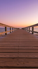 A serene wooden boardwalk leading to a tranquil beach at sunset. The sky is painted in soft pastel colors, with fluffy clouds scattered above. The calm ocean waves gently lap at the shore.