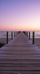 Obraz premium A serene wooden boardwalk leading to a tranquil beach at sunset. The sky is painted in soft pastel colors, with fluffy clouds scattered above. The calm ocean waves gently lap at the shore.