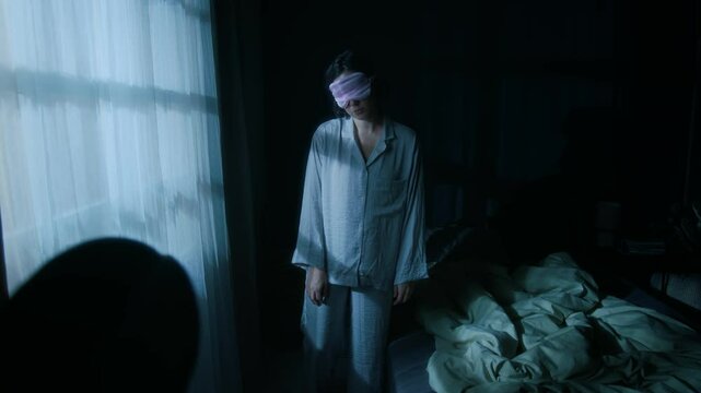 Young sleep-deprived woman in sleep mask and pajama sleepwalking at night in her dimly lit bedroom