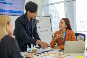 Three people are sitting at a table with a laptop open. One of them is laughing while the other two look on. The man in the middle is giving a presentation to the other two

