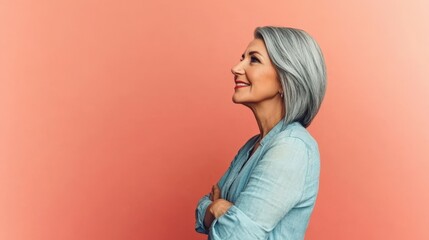 Fototapeta premium A confident older woman with silver hair smiles warmly while posing against a bright coral wall. She stands with her arms crossed, showcasing elegance and grace