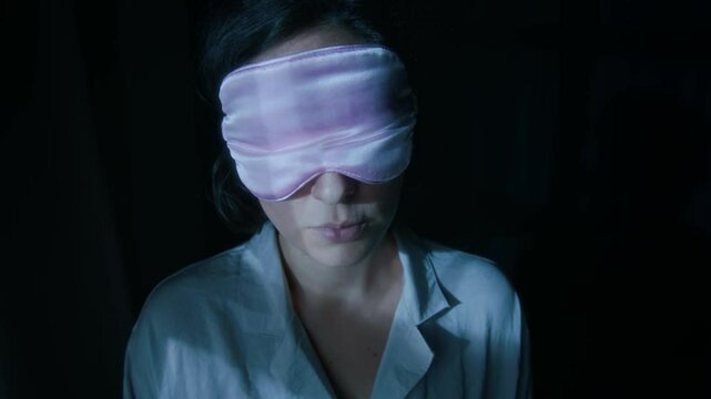 Close-up shot of young woman with dark short hair in pink sleep mask and pajama sleepwalking at night in dimly lit room