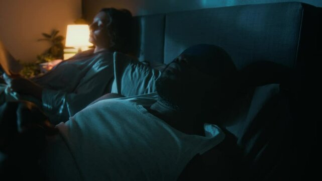 African American man, wearing sleep mask, turning off his bedside table lamp and falling asleep, while his Caucasian wife reading book, lying in bed with him
