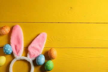 Beautiful decorated Easter eggs and bunny ears headband on yellow wooden table, flat lay. Space for text