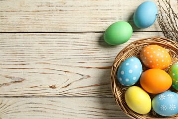 Beautiful decorated Easter eggs, wicker bowl and willow branches on light wooden table, flat lay. Space for text