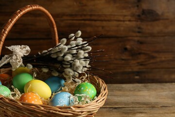 Beautiful decorated Easter eggs in wicker basket and willow branches on wooden table, closeup....