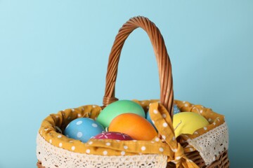 Beautiful decorated Easter eggs in wicker basket on light blue background, closeup