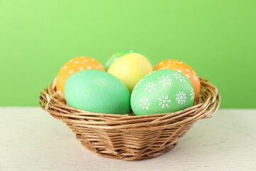 Many decorated Easter eggs in wicker basket on wooden table against light green background, closeup