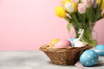 Easter eggs in wicker basket, bunny figure and beautiful tulips on grey table against pink background, closeup. Space for text