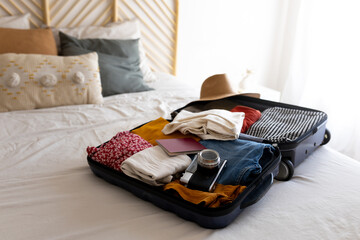 Open suitcase on the bed full of clothes and summer items: hat, sunglasses, swimsuit. Ready for...
